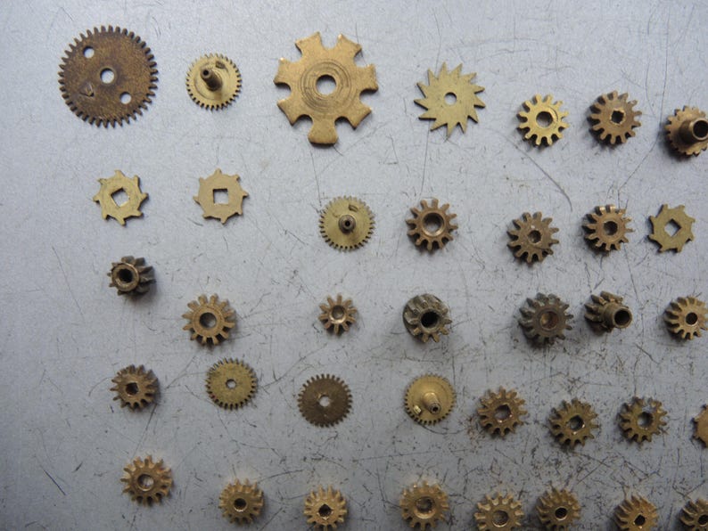 May include: A selection of small brass gears in various sizes and designs. The gears have different numbers of teeth and central holes. Some are circular, others more complex. The gears are scattered on a grey surface. Measurements are in centimetres.