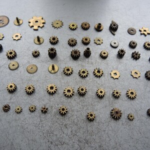 May include: A selection of small, brass-coloured gears in different sizes and designs. The gears are laid out on a grey surface, highlighting their intricate details. Suitable for watchmaking or steampunk projects.