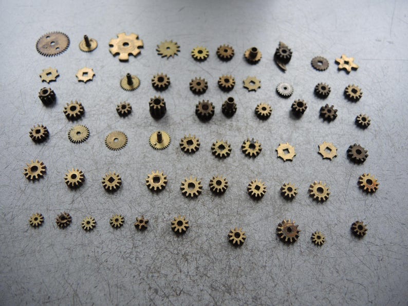 May include: A selection of small, brass-coloured gears in different sizes and designs. The gears are laid out on a grey surface, highlighting their intricate details. Suitable for watchmaking or steampunk projects.