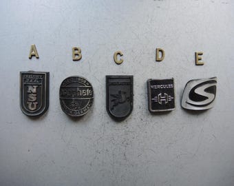 Könnte beinhalten: Fünf Vintage-Metall-Embleme mit unterschiedlichen Designs und Texten werden gezeigt. Die Embleme, in Formen wie Schilden und Ovalen, weisen geprägte Schriftzüge und Logos auf. Die Farbpalette ist hauptsächlich metallisch silber und schwarz, wobei jedes Emblem einzigartige Details aufweist.