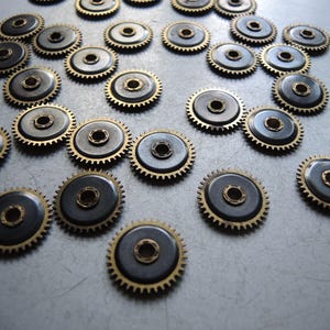 May include: A collection of small, metallic watch gears. Each gear has a dark gray center with a gold-colored toothed outer ring and a central hole. The gears are scattered across a light gray surface.