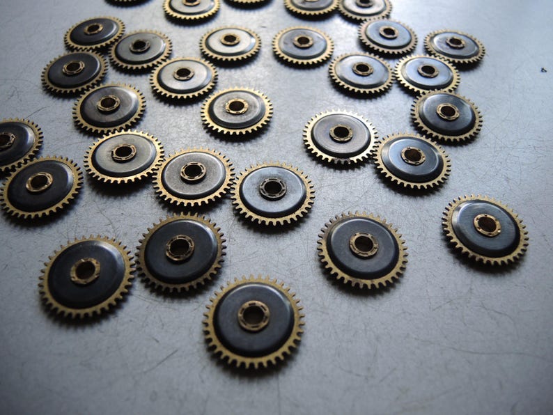 May include: A collection of small, metallic watch gears. Each gear has a dark gray center with a gold-colored toothed outer ring and a central hole. The gears are scattered across a light gray surface.