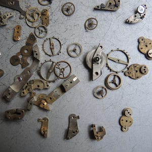 K&ouml;nnte beinhalten: Eine Sammlung von Vintage-Uhrenteilen, darunter Zahnr&auml;der, R&auml;der und Hebel, auf einer grauen Oberfl&auml;che. Die Metallkomponenten weisen Gebrauchsspuren auf, was darauf hindeutet, dass sie antik oder geborgen sind.