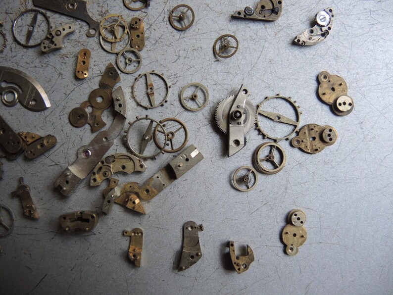 K&ouml;nnte beinhalten: Eine Sammlung von Vintage-Uhrenteilen, darunter Zahnr&auml;der, R&auml;der und Hebel, auf einer grauen Oberfl&auml;che. Die Metallkomponenten weisen Gebrauchsspuren auf, was darauf hindeutet, dass sie antik oder geborgen sind.