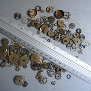 May include: A collection of vintage watch gears and components in various sizes and shapes, scattered on a gray surface. A metal ruler with inch markings is positioned diagonally across the gears. The gears are primarily brass and silver in color.