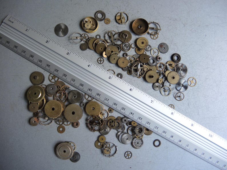 May include: A collection of vintage watch gears and components in various sizes and shapes, scattered on a gray surface. A metal ruler with inch markings is positioned diagonally across the gears. The gears are primarily brass and silver in color.