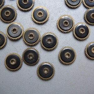 May include: A collection of small, brass-colored gears with black centers. The gears have a circular design with teeth around the edges and a central hole. The gears are scattered on a gray surface.