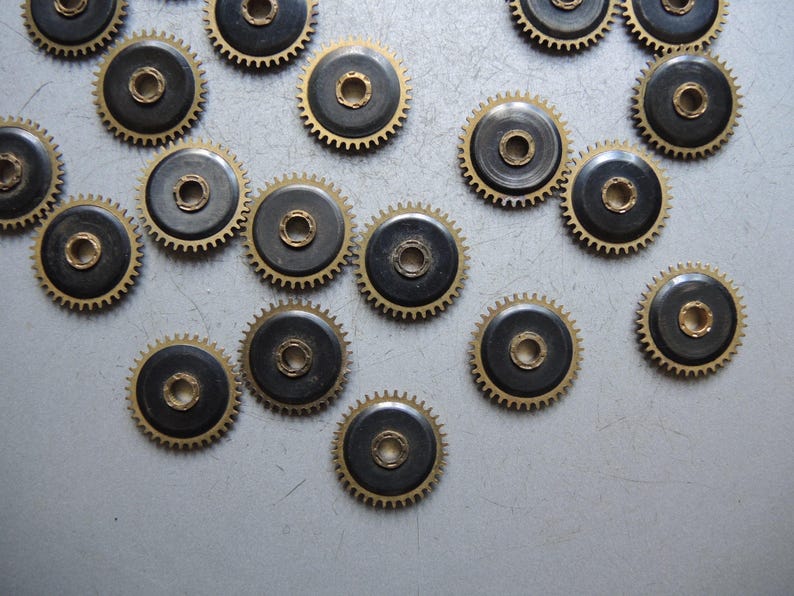 May include: A collection of small, brass-colored gears with black centers. The gears have a circular design with teeth around the edges and a central hole. The gears are scattered on a gray surface.