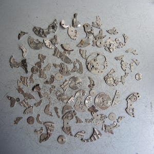 Pode incluir: Uma coleção de peças de movimento de relógio em metal prateado, incluindo engrenagens, placas, pontes e pequenos parafusos, espalhados numa disposição circular. Estes componentes mecânicos apresentam recortes intrincados e formas circulares.