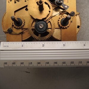 May include: Close-up of a clock mechanism with brass gears and components. A metal ruler with inch markings is placed across the mechanism. The ruler measures approximately 4 inches in length.