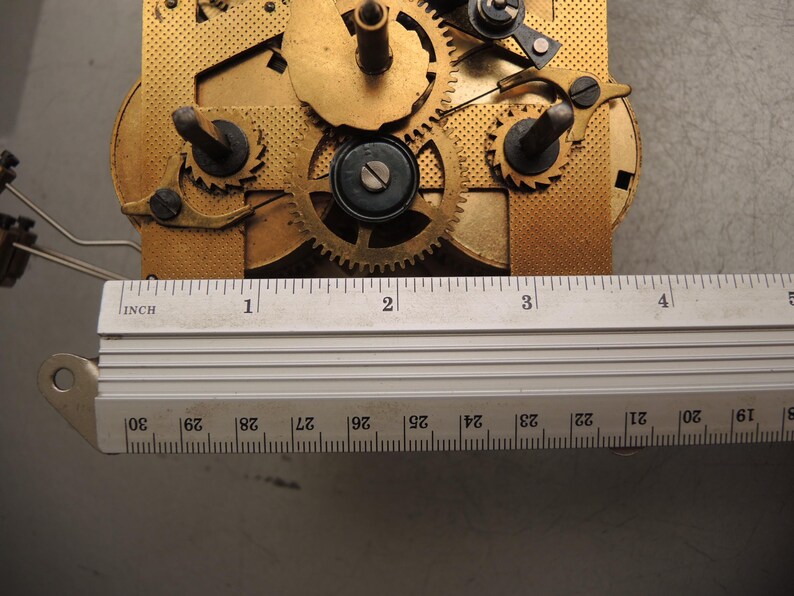 May include: Close-up of a clock mechanism with brass gears and components. A metal ruler with inch markings is placed across the mechanism. The ruler measures approximately 4 inches in length.