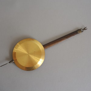 Puede incluir: Un péndulo de reloj de latón con un disco circular y una varilla de madera. El disco es de color dorado con un acabado cepillado. La varilla es marrón y termina en un extremo metálico. El péndulo es para un reloj.