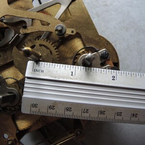 May include: Close-up of a clock mechanism with brass gears and components. A metal ruler is placed across the mechanism, showing measurements in inches. The gears are intricate and detailed, with a focus on the mechanical workings.