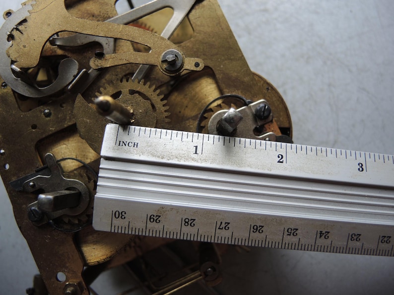 May include: Close-up of a clock mechanism with brass gears and components. A metal ruler is placed across the mechanism, showing measurements in inches. The gears are intricate and detailed, with a focus on the mechanical workings.