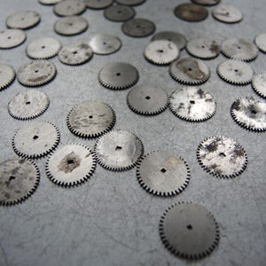 May include: A collection of small, silver-colored watch gears with toothed edges and square center holes. Some gears show signs of wear and discoloration, scattered across a gray surface. These vintage watch parts are ideal for repair or crafting.
