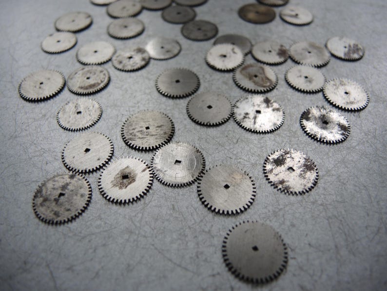 May include: A collection of small, silver-colored watch gears with toothed edges and square center holes. Some gears show signs of wear and discoloration, scattered across a gray surface. These vintage watch parts are ideal for repair or crafting.