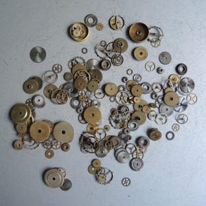 May include: A collection of various watch gears and components in shades of gold and silver, scattered on a light gray surface. The gears vary in size and design, showcasing intricate mechanical details.