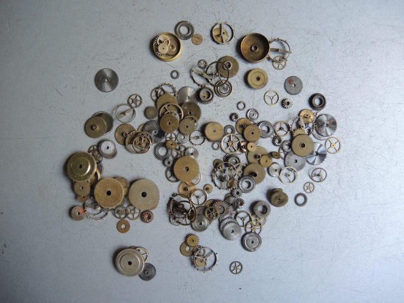 May include: A collection of various watch gears and components in shades of gold and silver, scattered on a light gray surface. The gears vary in size and design, showcasing intricate mechanical details.