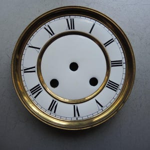 Könnte beinhalten: Ein Vintage-Zifferblatt mit weißem Zentrum und einem goldfarbenen Metallrahmen. Schwarze römische Ziffern markieren die Stunden. Das Zifferblatt hat drei schwarze Löcher in der Mitte.