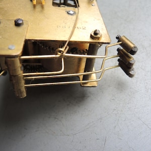 May include: Close-up of a vintage clock mechanism with brass components. The gears, levers, and other internal parts are visible. The metal has a golden hue, and the overall design is intricate and detailed.