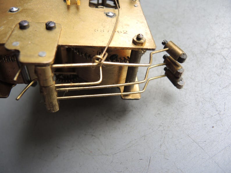 May include: Close-up of a vintage clock mechanism with brass components. The gears, levers, and other internal parts are visible. The metal has a golden hue, and the overall design is intricate and detailed.