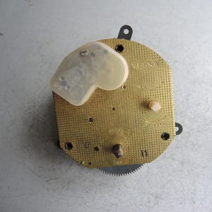 Op de afbeelding: Een uurwerk met een goudkleurige, getextureerde voorkant. Het uurwerk heeft een doorzichtige plastic afdekking en verschillende kleine tandwielen en componenten. Het woord "VESNA" is zichtbaar op de voorkant, samen met andere markeringen.