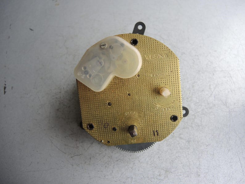 Op de afbeelding: Een uurwerk met een goudkleurige, getextureerde voorkant. Het uurwerk heeft een doorzichtige plastic afdekking en verschillende kleine tandwielen en componenten. Het woord "VESNA" is zichtbaar op de voorkant, samen met andere markeringen.