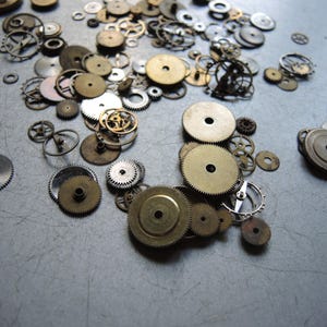 May include: A collection of various watch gears and cogs in different sizes and colors, including gold, silver, and bronze. The gears are scattered across a gray surface, showcasing intricate mechanical details.