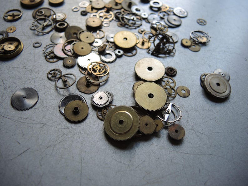 May include: A collection of various watch gears and cogs in different sizes and colors, including gold, silver, and bronze. The gears are scattered across a gray surface, showcasing intricate mechanical details.