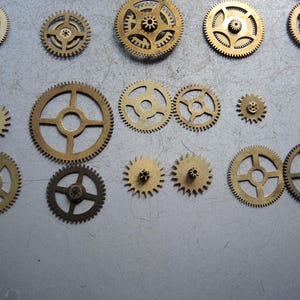 May include: A collection of various sized brass gears, arranged on a gray surface. The gears have intricate teeth and central holes, showcasing mechanical components. The gears vary in size and design, suitable for steampunk or industrial projects.