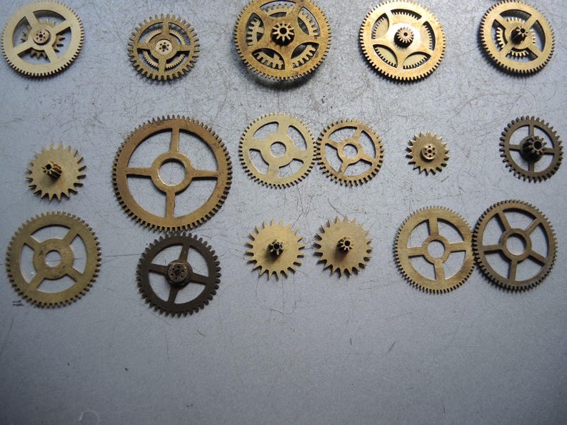 May include: A collection of various sized brass gears, arranged on a gray surface. The gears have intricate teeth and central holes, showcasing mechanical components. The gears vary in size and design, suitable for steampunk or industrial projects.