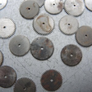 May include: A collection of small, circular metal gears with toothed edges and a central hole. The gears vary in color from silver to brown, showing signs of age and wear. These components are likely from a clock or watch.