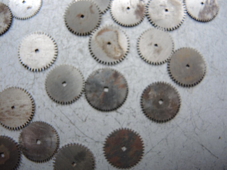 May include: A collection of small, circular metal gears with toothed edges and a central hole. The gears vary in color from silver to brown, showing signs of age and wear. These components are likely from a clock or watch.