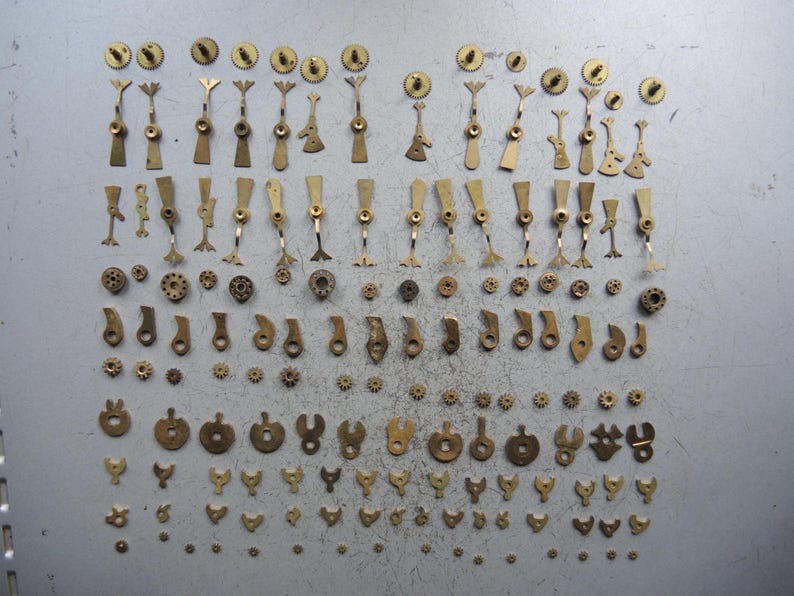 May include: A collection of vintage watch parts, including gears, levers, and other small components. The parts are made of brass and arranged on a gray surface. The image showcases a variety of shapes and sizes, ideal for watch repair or crafting.