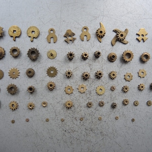 Puede incluir: Una colección de pequeños engranajes de latón de varias formas y tamaños. Los engranajes están dispuestos sobre una superficie gris, mostrando una variedad de ruedas dentadas, piñones y otros componentes mecánicos. Los engranajes varían desde diseños circulares simples hasta formas más complejas.