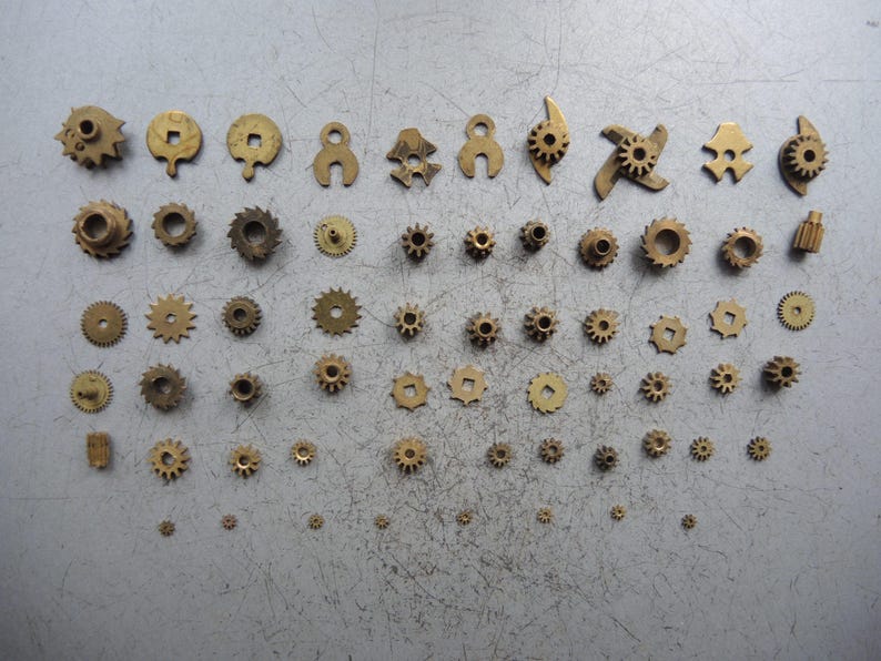 May include: A collection of small, brass gears of various shapes and sizes. The gears are arranged on a gray surface, showcasing a variety of toothed wheels, pinions, and other mechanical components. The gears range from simple circular designs to more complex shapes.