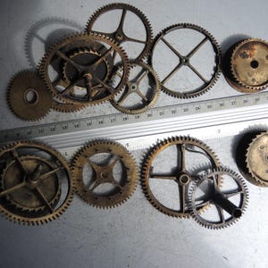 May include: A collection of various sized antique brass gears, some with visible teeth, arranged on a metallic surface. A metal ruler with inch markings is placed across the gears. The gears range from approximately 1 inch to 3 inches in diameter.