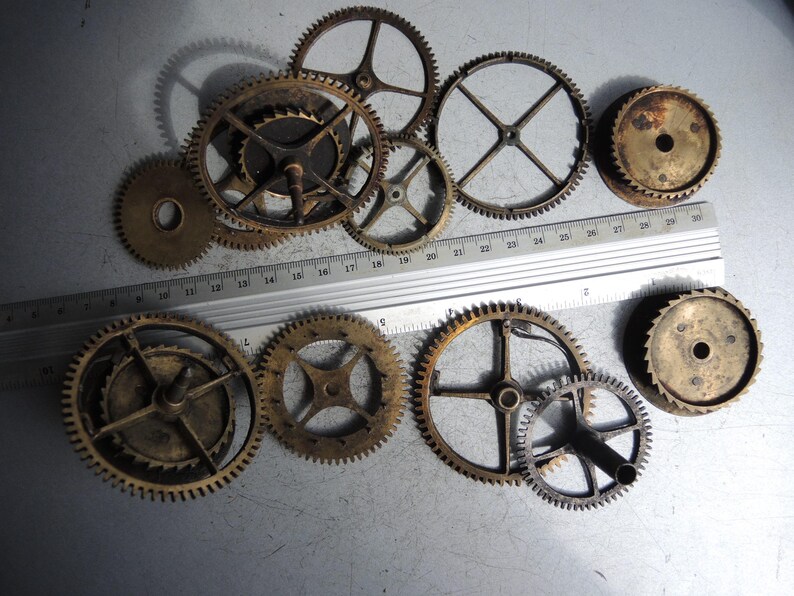 May include: A collection of various sized antique brass gears, some with visible teeth, arranged on a metallic surface. A metal ruler with inch markings is placed across the gears. The gears range from approximately 1 inch to 3 inches in diameter.