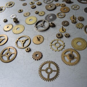 May include: A collection of various brass and silver-toned gears of different sizes and designs scattered on a gray surface. The gears include cogwheels, sprockets, and other mechanical components, suitable for watchmaking or steampunk projects.