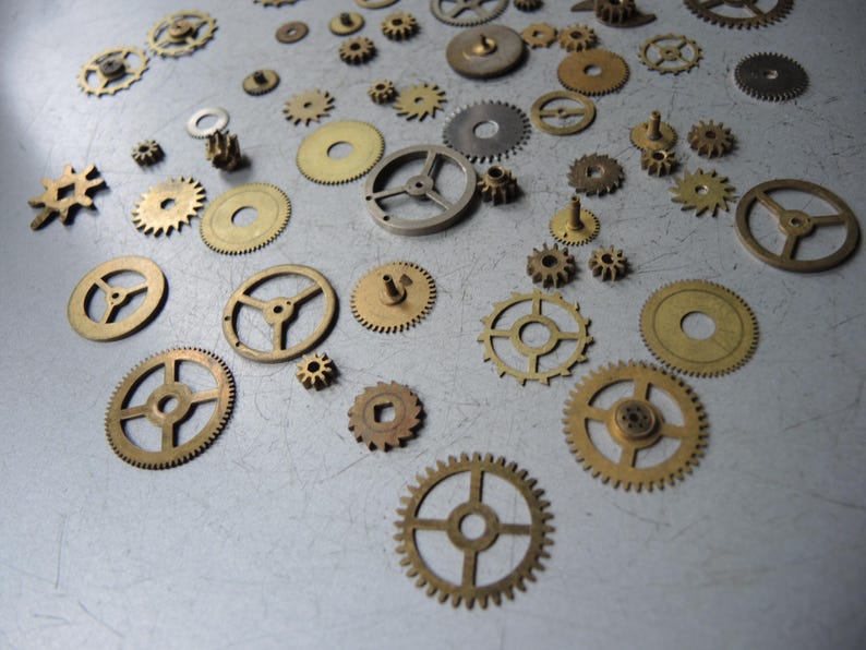 May include: A collection of various brass and silver-toned gears of different sizes and designs scattered on a gray surface. The gears include cogwheels, sprockets, and other mechanical components, suitable for watchmaking or steampunk projects.