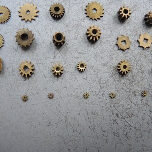 May include: A collection of small, brass gears of various sizes and designs, including spur gears and a worm gear. The gears are arranged on a gray, scratched surface, showcasing their intricate details and mechanical nature.