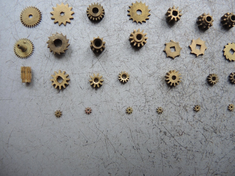 May include: A collection of small, brass gears of various sizes and designs, including spur gears and a worm gear. The gears are arranged on a gray, scratched surface, showcasing their intricate details and mechanical nature.