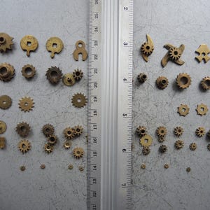 May include: A collection of small, brass gears and mechanical components of various shapes and sizes. The gears are arranged on a gray surface next to a ruler marked in inches, showcasing their dimensions.