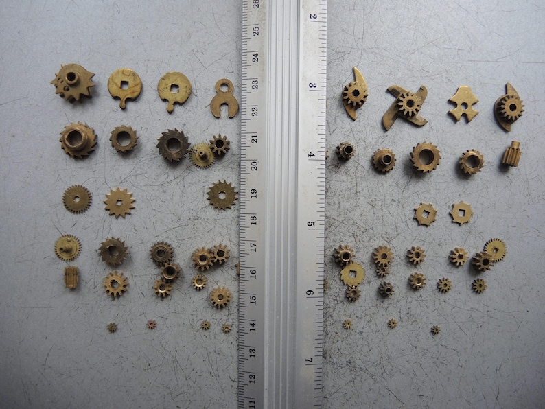 May include: A collection of small, brass gears and mechanical components of various shapes and sizes. The gears are arranged on a gray surface next to a ruler marked in inches, showcasing their dimensions.