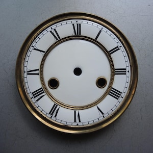 Könnte beinhalten: Ein Vintage-Zifferblatt mit weißem Hintergrund und schwarzen römischen Ziffern. Das Zifferblatt hat einen goldfarbenen Metallrahmen und zwei kleine Löcher in der Nähe der Mitte. Das Zifferblatt hat eine kreisförmige Form.