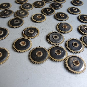 May include: A collection of small, circular gears with a brass-colored outer ring and a dark center. The gears are scattered across a light-colored surface, showcasing their intricate design and mechanical nature.