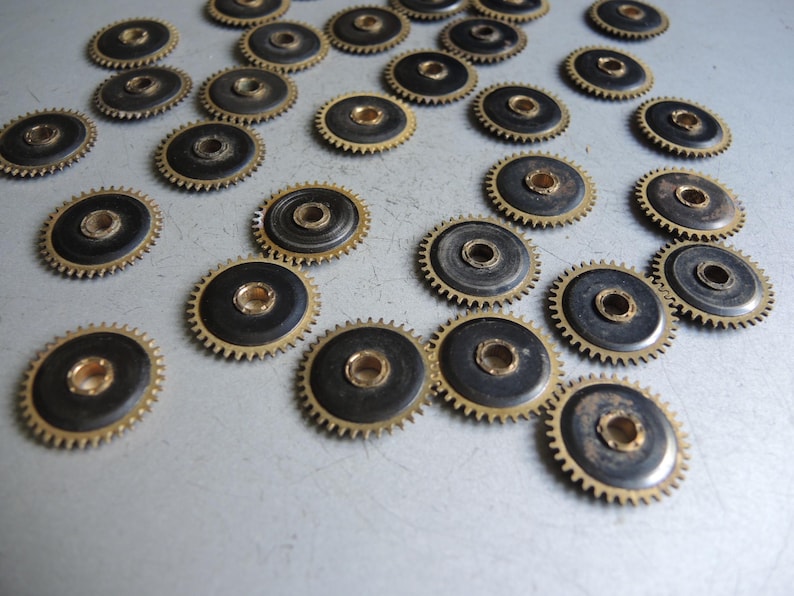 May include: A collection of small, circular gears with a brass-colored outer ring and a dark center. The gears are scattered across a light-colored surface, showcasing their intricate design and mechanical nature.