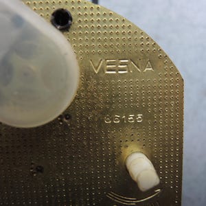 Op de afbeelding: Close-up van een messingkleurig uurwerkonderdeel. Het woord "VESNA" is bovenaan gegraveerd, met het nummer "86155" eronder. Een doorzichtig plastic onderdeel en een witte plastic knop zijn ook zichtbaar.