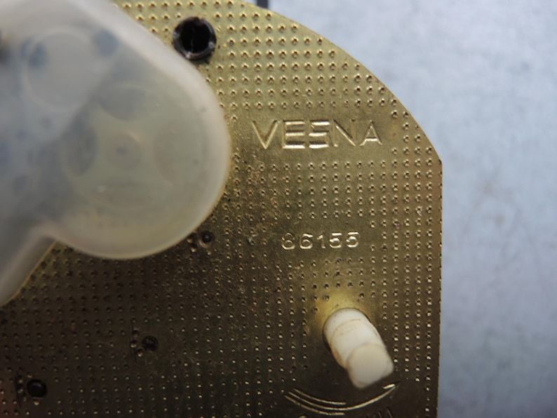 Op de afbeelding: Close-up van een messingkleurig uurwerkonderdeel. Het woord "VESNA" is bovenaan gegraveerd, met het nummer "86155" eronder. Een doorzichtig plastic onderdeel en een witte plastic knop zijn ook zichtbaar.