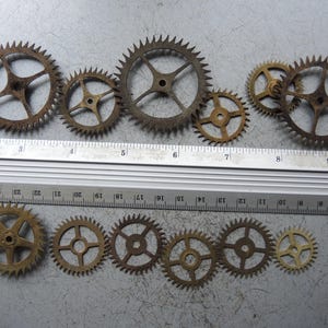 May include: A collection of antique brass gears of varying sizes, arranged on a gray surface. The gears have a weathered, aged appearance, with visible rust and discoloration. A ruler is placed next to the gears for scale, showing measurements in centimetres.